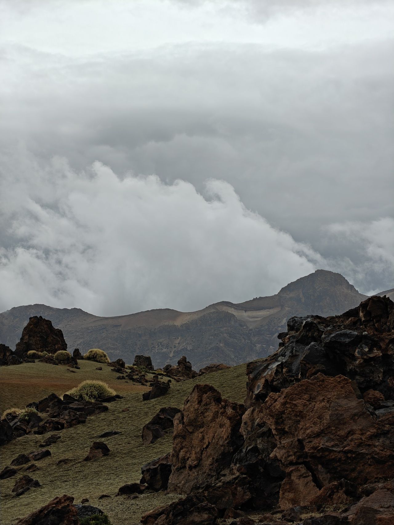 Wolken quellen über den Kraterrand, davor rostrote Lavafelsen und grünlicher Bimsboden
